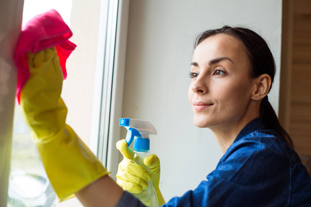 Beautiful young woman using a duster and a spray while cleaning windows in the house - Foto, afbeelding