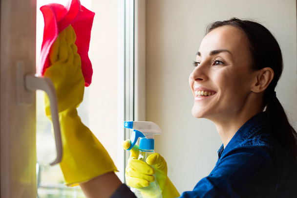 Beautiful young woman using a duster and a spray while cleaning windows in the house - Foto, immagini