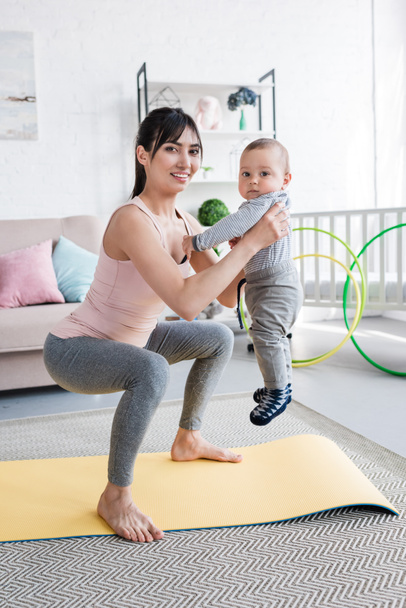 young sportive mother carrying her little child while doing squats - Foto, Bild