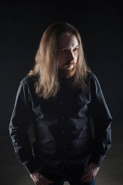Portrait of man with long hair and beard in black clothes on dark background - Fotografie, Obrázek