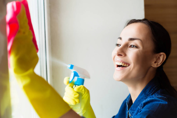 Hermosa mujer joven usando un plumero y un aerosol mientras limpia ventanas en la casa
 - Foto, Imagen