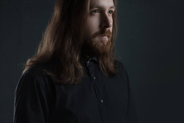 Portrait of man with long hair and beard in black clothes on dark background - Valokuva, kuva