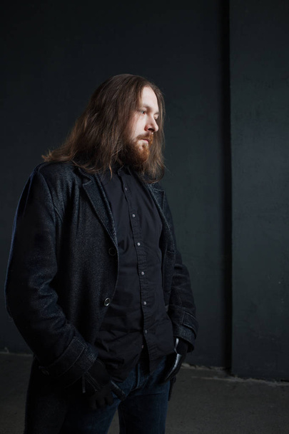Portrait of man with long hair and beard in black clothes on dark background - Φωτογραφία, εικόνα