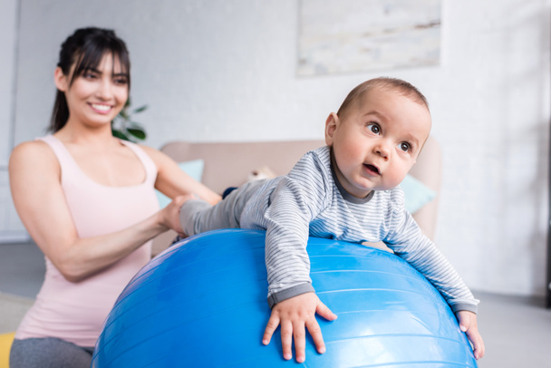 junge glückliche Mutter spielt mit kleinem Kind, während er zu Hause auf einem Ball liegt - Foto, Bild