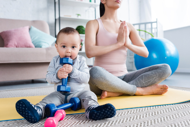 junge Mutter meditiert in Lotus-Pose, während ihr kleines Kind zu Hause eine Hantel hält - Foto, Bild