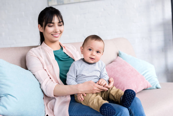 молодая счастливая мать с маленьким ребенком, сидящим дома на диване
 - Фото, изображение