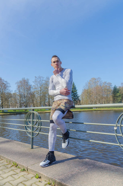 attractive man in white sportswear sitting on a railing by the river - Photo, Image