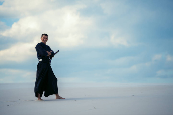 Confident smiling man practicing martial arts posing in the desert, in traditional Japanese clothes, with a Japanese sword - a katana. - Фото, изображение