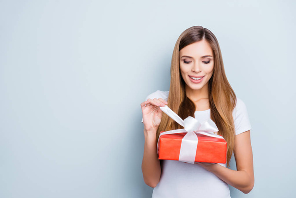 Portrait with copy space empty place of positive cute girl in white t-shirt opening gift box in red package with white ribbon isolated on grey background - 写真・画像