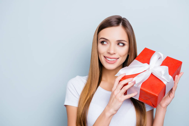 Portrait with copyspace empty place of nice gorgeous girl holding gift box in red package with white ribbon near ear want to know what inside isolated on grey background - Foto, afbeelding