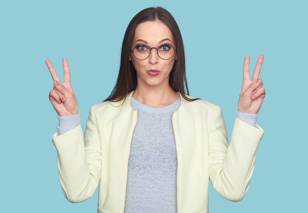 woman showing peace sign. Cheerful pretty girl wearing glasses isolated on bright blue background - Photo, Image