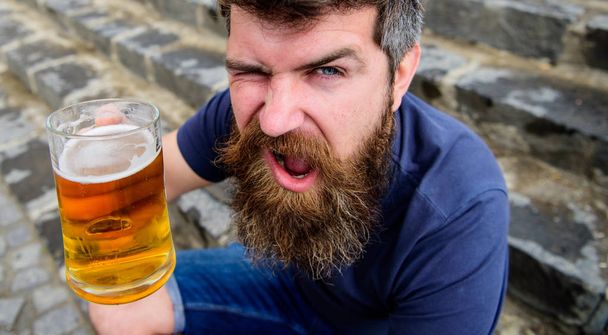 Celebration concept. Man with beard and mustache holds glass with beer while sits on stone stairs, defocused. Hipster on cheerful winking face drinks beer outdoor. Guy having rest with draught beer - Foto, Imagen