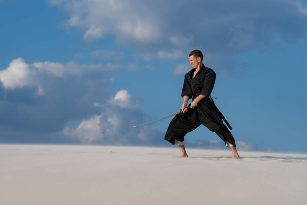Resolute man, in traditional clothes, is practicing Japanese martial art - iaido in the desert with a Japanese sword - a katana during sunset on the background of evening sky. Side view. - Foto, Imagen