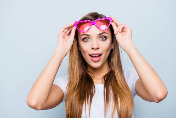 Portrait with copy space, empty place of impressed wondered girl raised her pink glasses in heart shape with wide open eyes mouth isolated on grey background - Foto, immagini