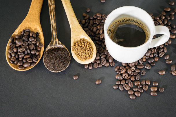 heißer Kaffee in weißem Keramikglas und Kaffeebohnen, Kaffeepulver und Instantkaffee werden in einem Holzlöffel auf einer schwarzen Tischplatte platziert. - Foto, Bild