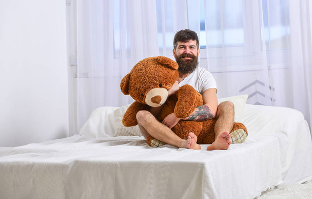 Guy on cheerful face hugs giant teddy bear. Man sits on bed and hugs big toy, white curtains on background. Macho with beard and mustache cuddling with plush toy on birthday morning. Birthday concept - Photo, Image