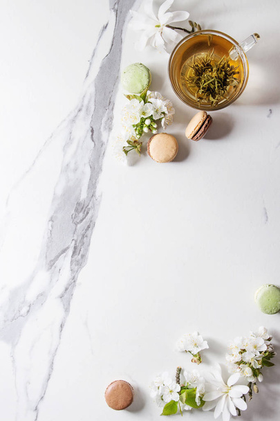 Glass cup of hot green tea with french dessert macaroons, spring flowers white magnolia and cherry blooming branches over white marble texture background. Top view, copy space. - Photo, image