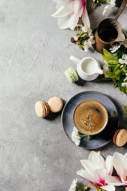 Blue cup of black espresso coffee french dessert macaroons, cream and spring flowers magnolia, blooming cherry branches over grey texture background. Top view, space. Spring greeting card - Foto, Imagem
