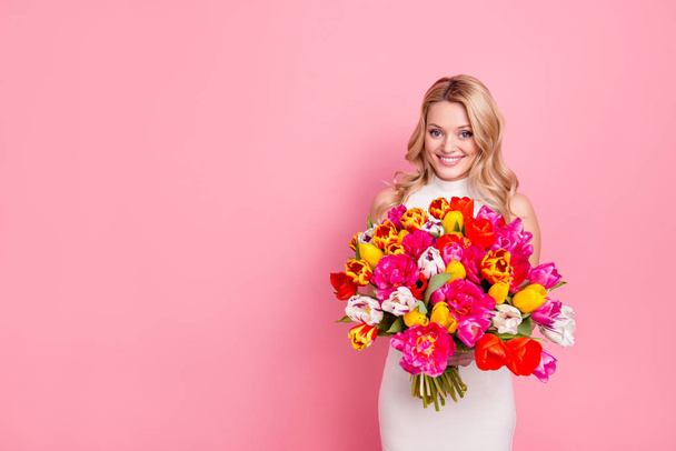Portrait with copyspace, empty, place of gorgeous positive woman in white dress having big bouquet of tulips in hands looking at camera isolated on pink background - Foto, Bild