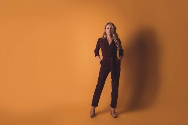 Full size, fullbody portrait of  arrogant stylish stunning woman in overalls holding two hands in pockets looking at camera isolated on dark yellow background with copy space - Foto, imagen