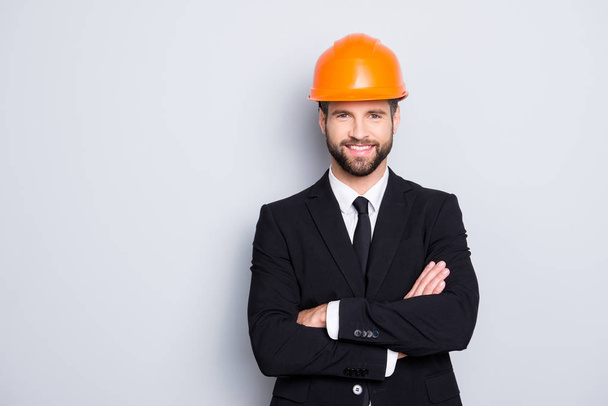 Portrait of attractive young stylish investor with stubble in orange hard hat and black tux, tuxedo with tie, looking at camera, isolated on grey background, having his arms crossed - Фото, изображение
