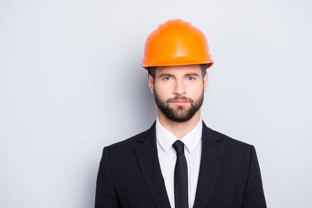 Portrait of concentrated, thoughtful architector with stubble in orange safety helmet, hardhat and black tux with tie, looking at camera, isolated on grey background - Photo, Image