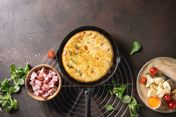 Spanish potato omelette tortilla with bacon served in cast-iron pan on cooling rack with ingredients above over over dark brown texture background. Top view, space. - 写真・画像