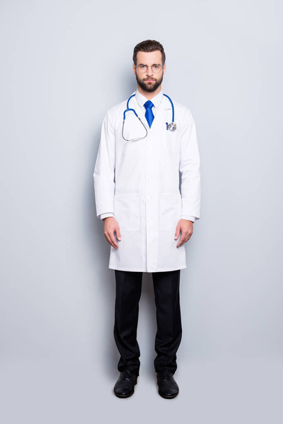 Full size portrait of attractive, concentrated virile harsh doc with beard in white lab coat, tie and stethoscope on his neck, looking at camera, isolated on grey background - Φωτογραφία, εικόνα