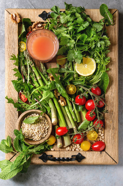 Variety of vegetarian healthy eating food ingredients. Green asparagus, herbs, tomatoes, nuts, wheat corns, dandelion leaves, glass of juice on wood tray over grey texture background. Top view, space. - Foto, afbeelding