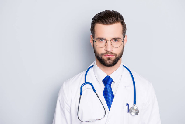Portrait of trendy modern doc with stubble and modern hairstyle in white lab coat, tie and stethoscope on his neck, looking at camera, isolated on grey background - Foto, Bild