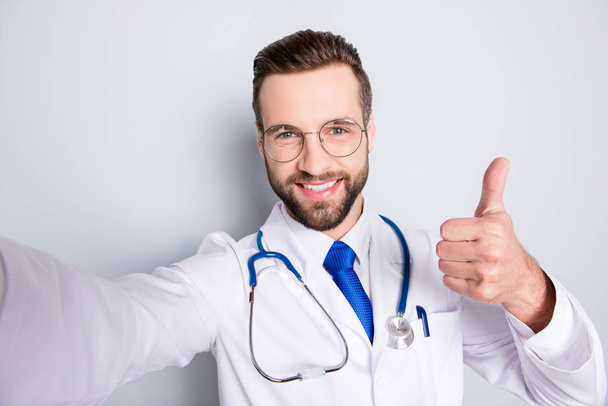 Self portrait of positive trendy doc in white outfit with tie and bristle having stethoscope on his neck shooting selfie showing thumb up, like sign with finger, hand over grey background - Foto, Bild