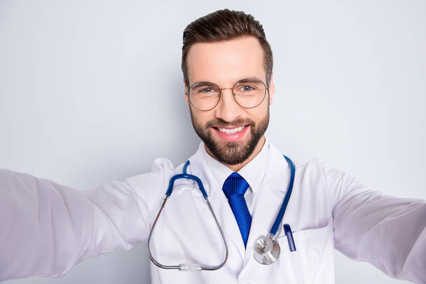 Self portrait of joyful cheerful doc in white outfit with tie and stubble having stethoscope on his neck shooting selfie with two arms, isolated over grey background - Photo, Image