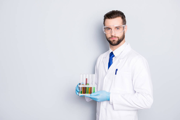 Portrait with copy space, empty place of attractive stylish  scientist with bristle in white lab coat, tie holding test tubes with multi-colored liquid, looking at camera over grey background - Photo, Image