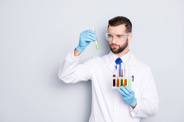 Portrait of busy concentrated scientist with stubble in white lab coat, gloves analysing, looking at  test tubes with multi-colored liquid in his arm, isolated on grey background - Fotó, kép