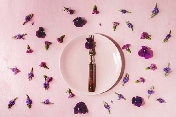 Variety of purple edible flowers for dish decorating with empty ceramic plate and fork over pink pastel background. Top view, space. - Fotografie, Obrázek