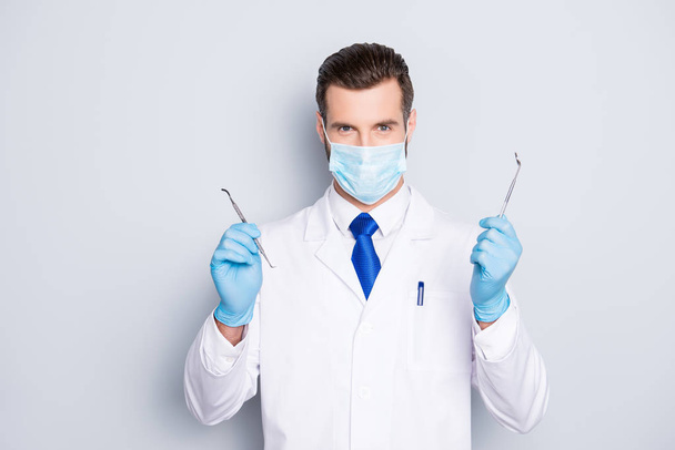 Portrait of successful handsome dentist in white lab coat with blue tie having, showing, holding tools for teeth treatment with hands, looking at camera, isolated on grey background - Фото, зображення