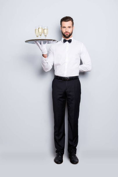 Full size fullbody portrait of concentrated man in classic white shirt and black bowtie holding hand behind the back and tray with three glasses of sparkling wine, isolated on grey background - Photo, Image