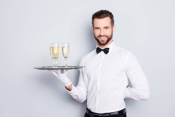 Portrait of cheerful friendly man in classic white shirt and black bow holding hand behind the back and tray with three glasses of champaign, sparkling wine, isolated on grey background - Photo, image
