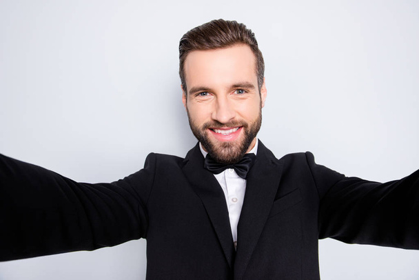 Self portrait of stylish cheerful artist in black suit with bowtie, shooting selfie on front camera of smart phone with two arms, having video-call, isolated on grey background - Φωτογραφία, εικόνα
