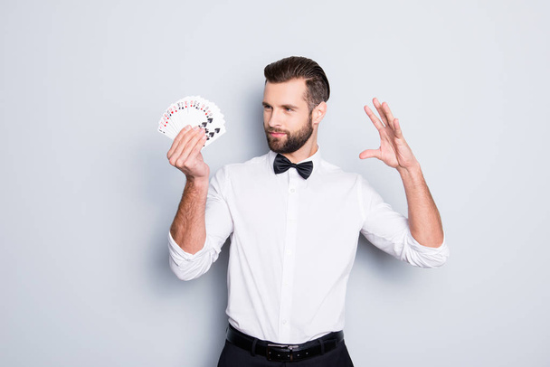 Portrait of cunning foxy magician with modern hairstyle and stubble in white shirt with bowtie holding set of cards in hand, making trick, focus, isolated on grey background - Fotografie, Obrázek