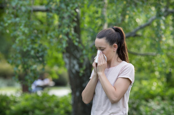 Молодая красивая женщина сморкается в парке с деревьями на заднем плане. Аллергия на пыльцу весной
 - Фото, изображение