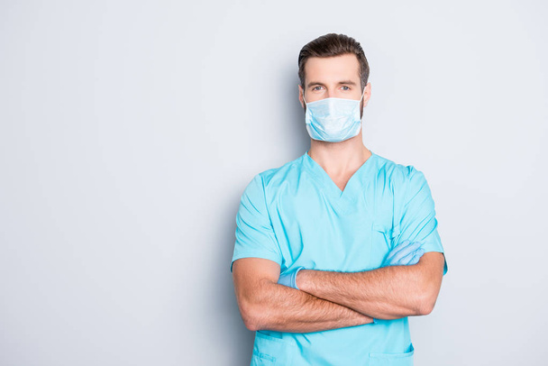 Portrait with copyspace, empty place of attractive handsome man in sterile mask and blue lab uniform, having his arms crossed, looking at camera isolated on grey background - Foto, afbeelding