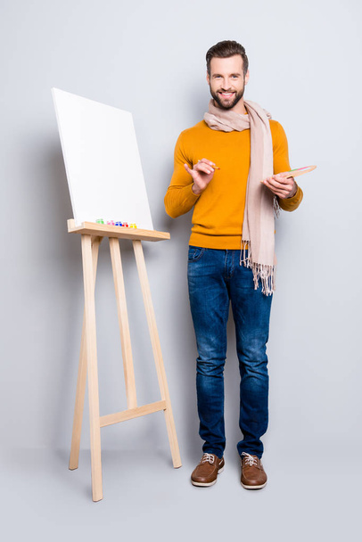 Full size body portrait of joyful trendy artist with scarf around neck, hairstyle, in jeans, sweater, holding colorful palette and brushes in hands, isolated on grey background - Photo, image