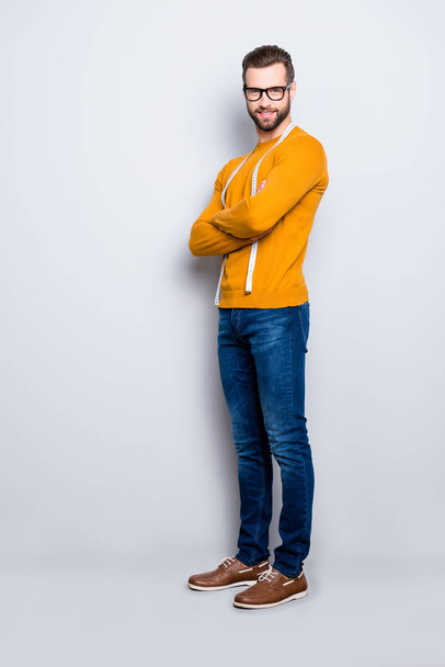 Full size body portrait of cheerful joyful half-turned dressmaker with centimeter on neck in pullover, jeans holding arms crossed, looking at camera isolated on grey background - Fotó, kép