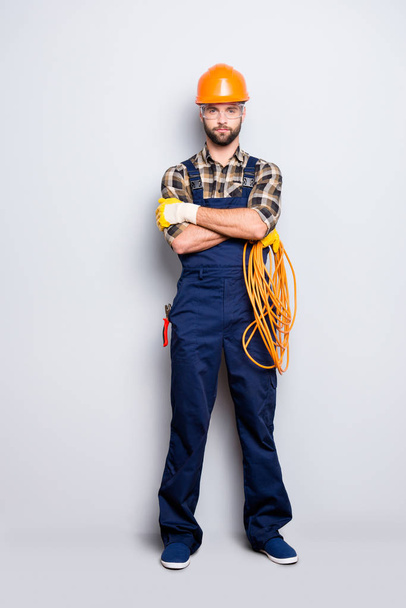 Full size body portrait of virile brutal electrician with bristle in overall, shirt, holding rolled cable in hand, having his arms crossed, looking at camera, isolated on grey background - Fotó, kép