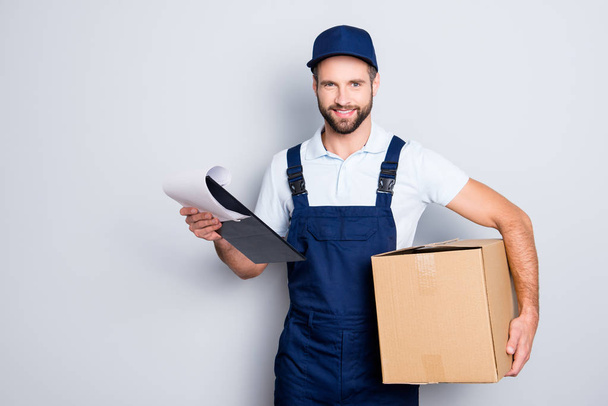 Portrait of cheerful positive deliver in blue uniform carrying having box and clipboard in hands, looking at camera, isolated on grey background - 写真・画像