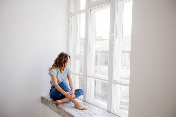 Beautiful girl by the window on a sunny day, dressed in jeans and a blue T-shirt. - Foto, Bild