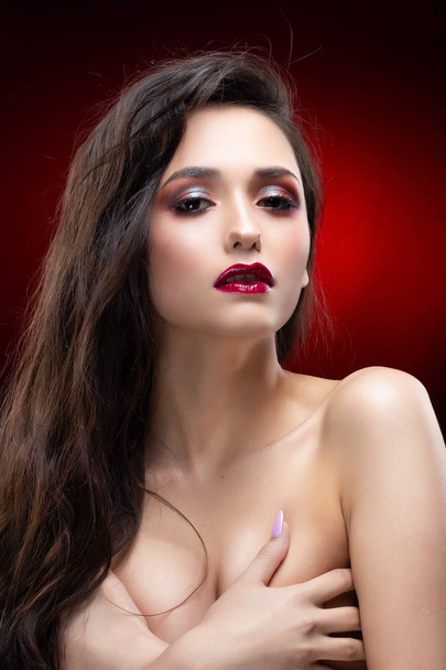Portrait of a beautiful glamorous brunette girl sensually covers her naked breast with her hands. Shimmering makeup, red lips. Healthy smooth skin. Black and red background. Copy space. - Fotografie, Obrázek
