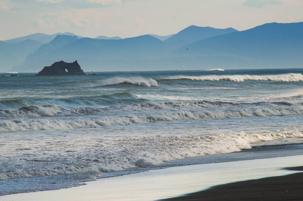 Halaktyr beach. Kamchatka. Russian federation. Dark almost black color sand beach of Pacific ocean. Stone mountains and yellow grass are on a background. Light blue sky - Photo, Image
