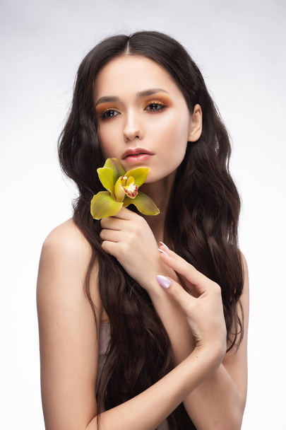 A beautiful brunette nude shoulders girl sensually holds a flower of an orchid in her hands. Nude make-up. Healthy smooth skin. Isolated on white background. Copy space. Commercial and advertising. - Foto, Imagem